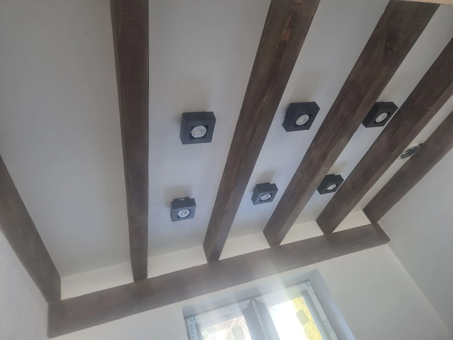 Zdjęcie przedstawia sufit z podwójną warstwą gipsu z ciemnymi belami drewnianymi oraz montowanymi oprawami oświetleniowy